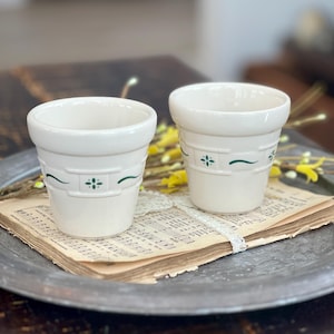 May include: Two off-white ceramic flower pots with green floral accents. The pots are on a metal tray atop vintage sheet music. The pots have a classic, simple design, perfect for small plants or herbs.