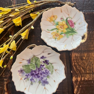 May include: Two leaf-shaped, white porcelain dishes with gold trim. One dish features a bouquet of yellow daffodils and a pink rose. The other dish displays a cluster of purple violets and green leaves. Both have a small handle.