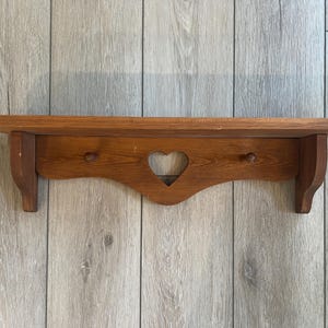 May include: A medium-toned wooden wall shelf featuring a flat top surface and a decorative back panel. The back panel includes a central heart-shaped cutout and two small wooden pegs positioned on either side for hanging items.