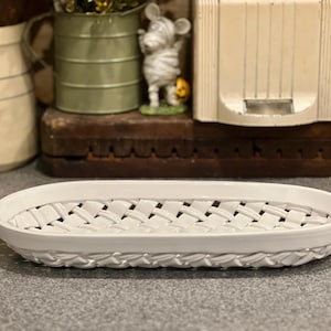 May include: White ceramic oval basket with a woven design. The basket has a raised, braided edge and a lattice pattern across the center. This decorative piece is suitable for serving or display.