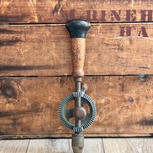 May include: A vintage hand drill with a wooden handle and a metal gear mechanism. The drill is in good condition and has a black painted handle.