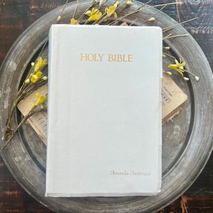 May include: A white hardcover Holy Bible with gold lettering on the front cover. The text reads HOLY BIBLE at the top and Amanda Antenucci at the bottom right. The book is positioned on a round metallic tray with yellow floral sprigs.