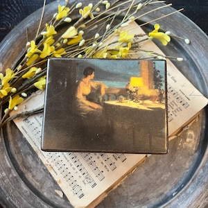 May include: A rectangular wooden box featuring a printed image of a person sitting at a table on a balcony overlooking a body of water at dusk, with a glowing lamp on the table. The box has a dark finish and a smooth, flat top surface.