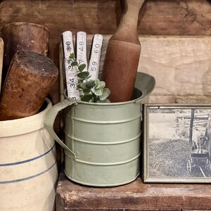 May include: A collection of vintage kitchen items. A light green metal pitcher holds a wooden rolling pin, measuring sticks, and greenery. A framed black and white photo sits next to the pitcher. Other items include a ceramic jar and wooden objects.