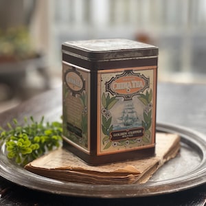 May include: A rectangular brown metal tea tin featuring a label with the text Choice China Tea and Golden Clipper Brand. The label displays a graphic of a sailing ship surrounded by green leaves and floral accents on a cream-colored background.