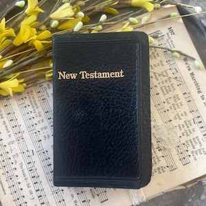May include: A small, black New Testament book with gold lettering, resting on vintage sheet music. The book's cover has a textured appearance. Yellow floral accents add a touch of color to the composition.