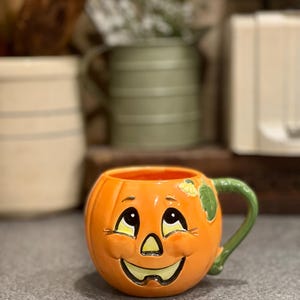 May include: Orange ceramic pumpkin-shaped mug with a smiling jack-o'-lantern face. The mug has a green handle and a yellow leaf detail. The interior is orange. A festive Halloween-themed item.