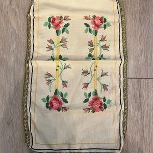 May include: A rectangular cream-coloured fabric piece featuring a cross-stitch pattern of two vertical yellow stripes flanked by pink rose blossoms and green leaves. The edges are finished with a decorative metallic gold and black braided trim.