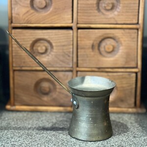 May include: A metallic coffee pot with a long, slender handle and a wide, flared rim. The body of the pot has a cylindrical shape that tapers slightly toward the center, featuring a weathered, tarnished finish with visible spots and discoloration.