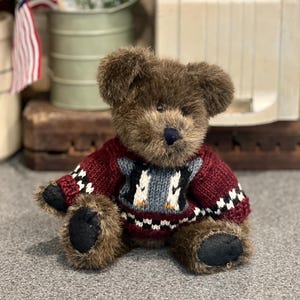 May include: A brown teddy bear wearing a burgundy sweater with a penguin design. The bear has a dark blue nose and black paw pads. The sweater has white and black accents. The bear is sitting on a gray surface.