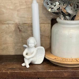 May include: A white ceramic angel figurine with folded wings sits on a wooden surface. A tall white candle stands behind the angel. A white ceramic jar with metal serving spoons is in the background.
