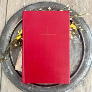 May include: A rectangular book with a vibrant red textured cover featuring a thin gold-coloured cross centred on the front. The book is positioned vertically, showcasing a clean, minimalist design with a solid colour finish and a vertical spine edge.