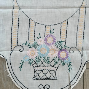 May include: A white fabric piece featuring an embroidered basket filled with purple, blue, and pink flowers with green leaves. The design includes black outlined scrollwork and vertical yellow stitched lines on a light-colored textile background.