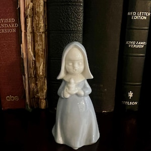 May include: A glossy porcelain figurine of a person wearing a light blue hooded robe and holding a small white candle. The figure has a smooth, rounded face and a simple, flowing garment that extends to the base, standing in front of dark books.