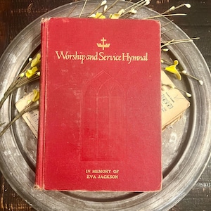 May include: A red hardcover book titled Worship and Service Hymnal with a gold cross symbol and an embossed gothic window design on the front cover. The text In Memory of Eva Jackson is printed in gold at the bottom of the red fabric cover.