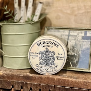 May include: A vintage tin of Burgess's Genuine Anchovy Paste for Toast Biscuit &c. The round tin has a cream-colored lid with text and a crest. A small green metal bucket and a framed black and white photo are in the background.