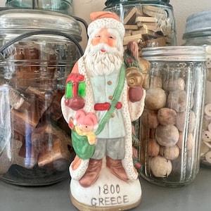 May include: A ceramic Santa Claus figurine wearing a white-trimmed coat, holding a green gift and a sack containing a small pink-hatted figure. The base is inscribed with the text 1800 GREECE. The figure features a white beard and hat.