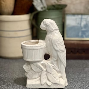 May include: A white ceramic parrot figurine with a small bucket-shaped planter. The parrot is perched on a branch with detailed leaves. The figurine is a decorative item, likely for a home or garden.