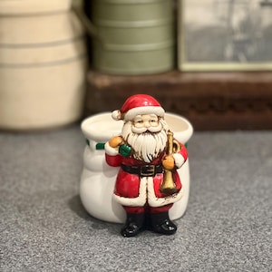 May include: A white ceramic planter featuring a three-dimensional Santa Claus figure on the front. Santa wears a red suit with white trim, a black belt, and black boots, holding a small green sack in one hand and a gold-colored trumpet in the other.