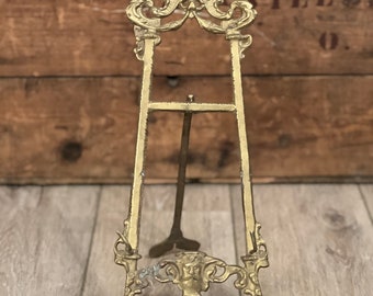 May include: A gold-toned metal easel with an ornate design featuring a face at the top and a decorative base. The easel is standing on a wooden surface.