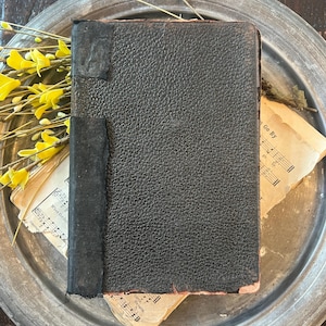 May include: A vintage, textured black book with a dark spine, resting on a silver-toned metal tray. Yellow flowers and aged sheet music surround the book. The book's cover shows wear and tear, suggesting age and use.