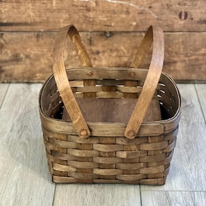 May include: A rectangular woven basket with a dark brown finish, featuring two arched wooden handles attached to the rim with copper-colored rivets. The basket has a flat, solid wooden base and a woven body with a consistent, tight grid pattern.