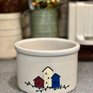 May include: A cream-colored ceramic pot with a rounded rim. The pot is decorated with three painted birdhouses in red, white, and blue, perched on a green vine. The birdhouses have black roofs. The pot is likely for decorative use.