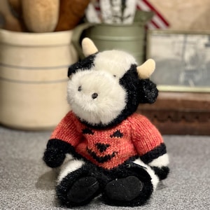 May include: A plush cow toy with black and white fur, featuring small tan horns and black plastic eyes. The cow is wearing a knitted orange sweater with a dark brown jack-o'-lantern face pattern on the front, including eyes and a jagged mouth.