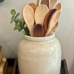 May include: A collection of various wooden spoons in different shades of brown, ranging from light tan to dark mahogany, are arranged inside a large, cylindrical, off-white ceramic crock, accompanied by a small sprig of green eucalyptus leaves.