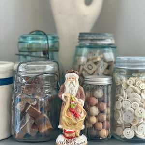 May include: A ceramic Santa Claus figurine with a sack of toys stands in front of several glass jars filled with various items. The jars are arranged on a gray surface. The figurine is marked "1897 Czechoslovakia".