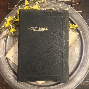 May include: A black, textured, hardcover Holy Bible Concordance with gold lettering on the front cover. The book is positioned on a round, metallic, silver-toned tray, accompanied by yellow floral sprigs and a piece of vintage sheet music.