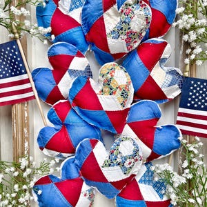 May include: A collection of patriotic heart-shaped ornaments in red, white, and blue patchwork fabric. The hearts are arranged in a wooden frame, with small American flags and white flowers. The overall theme is Americana.