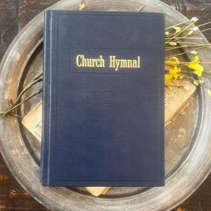 May include: A dark blue hardback book with the words Church Hymnal embossed in gold on the front cover. The book features a rectangular border design and a small circular emblem centred below the title text on the front cover surface.