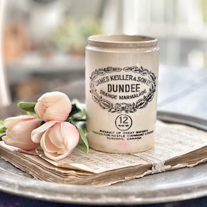 May include: A vintage white ceramic jar with black lettering that reads "JAMES KEILLER & SON DUNDEE ORANGE MARMALADE 12" and "PRODUCT OF GREAT BRITAIN PACKED FOR NESTLE (CANADA) LTD TORONTO, CANADA". The jar is sitting on a stack of old sheet music on a silver tray.