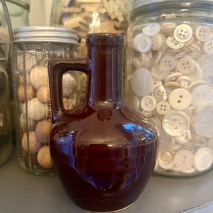 May include: A small, dark brown ceramic jug with a glossy finish, featuring a rounded body and a single side handle. The jug has a narrow neck and a flared rim, standing upright with a smooth, reflective surface that catches the light.
