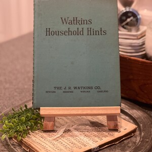 以下が含まれることがあります： 「Watkins Household Hints」というタイトルのヴィンテージグリーンの書籍。出版社は「The J.R. Watkins Co.」で、表紙には「Newark、Memphis、Winona、Oakland」の場所が印刷されています。書籍は木製イーゼルに飾られています。