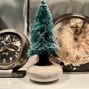 May include: A small, teal bottle brush Christmas tree with white frosted tips sits atop a cream-colored, ceramic base. Two vintage alarm clocks with black faces and silver casings are in the background, suggesting a holiday or seasonal theme.