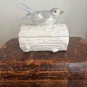 May include: A ceramic trinket box with a bird figurine on top. The bird is light gray with a brown beak, perched on a white box shaped like a log. The box is resting on a vintage, brown leather-bound book.