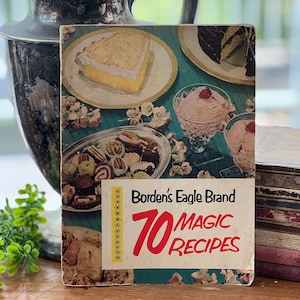 May include: Vintage cookbook cover with a turquoise background featuring a variety of desserts, including a lemon meringue pie, chocolate cake, and ice cream. The cover reads "Borden's Eagle Brand 70 Magic Recipes".