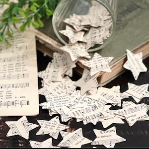 May include: A scattering of white paper stars with musical notes and text printed on them. The stars are scattered on a wooden surface and a book with a green cover. The text on the stars includes "Crown", "Almighty", "Pats in", "Manoak", "shalt", "Goo", "w Bridges.", "To", "ver me. My", "ny griefs Bet", "Up-", "Fran", "hip", "Ho-ly. Ho", "ly Thou art ho", "herbim and ser-a".