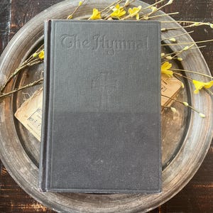 May include: A dark grey hardback book titled The Hymnal with an embossed Celtic cross on the front cover. The book is positioned on a round, weathered metallic tray decorated with thin, light brown branches featuring small yellow buds.