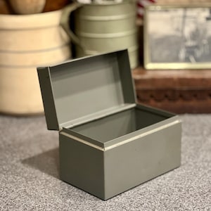 May include: A rectangular, olive-drab metal storage box with an open, hinged lid. The container features a simple, utilitarian design with clean lines and a smooth, matte finish, providing an open interior space for storage or organization.