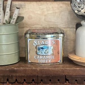 May include: A vintage Slades Caramel Toffy tin, rectangular in shape, with a label featuring "Crystal Spring" and "Caramel Toffy". The tin is brown and gold with a landscape illustration. A green container and a white ceramic jar are also visible.