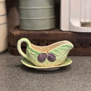 May include: A green ceramic gravy boat with a leaf design and two purple plums. The boat is on a matching saucer.