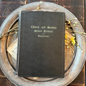 May include: A black hardcover book titled Church and Sunday School Hymnal with Supplement in gold lettering on the front cover. The book features a rectangular embossed border and shows signs of wear along the spine and edges of the cover.