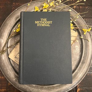 May include: A dark blue hardback book with the title THE METHODIST HYMNAL embossed in gold lettering on the front cover. The book features a textured, fabric-like finish and is positioned vertically, showing the title clearly in the upper centre.