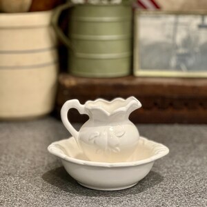 May include: A white ceramic washbasin set consisting of a small pitcher with a scalloped rim and embossed fruit detailing, paired with a matching shallow bowl featuring a scalloped edge. The set has a glossy finish and a classic rounded shape.