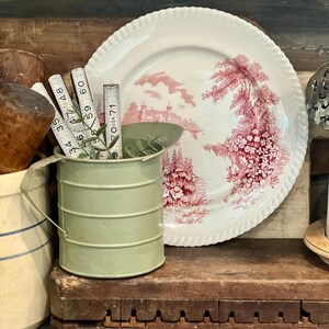 May include: A vintage-style still life featuring a white plate with a red floral design, a small green metal container, and a wooden ruler with measurements in inches. The plate has a scalloped edge. The scene evokes a rustic, antique aesthetic.