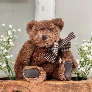 May include: A brown teddy bear with a black nose and dark brown paw pads. The bear has a checkered bow and is sitting on a wooden surface. White flowers are in the background.