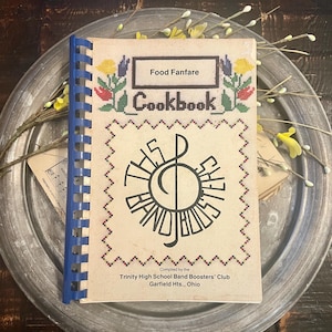 May include: A spiral-bound cookbook titled Food Fanfare Cookbook, featuring a cross-stitch style floral border and a central treble clef graphic with the text This Band Boosters. The cover includes the text Compiled by the Trinity High School Band Boosters' Club, Garfield Hts., Ohio.